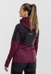 Fleece jacket with a maroon body and black padded section on shoulders. Features a hood and a logo on the back. Soft texture, long sleeves.