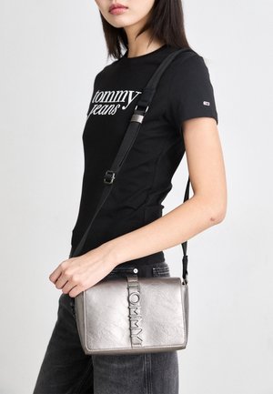 Kobieta w czarnej koszulce Tommy Jeans i ciemnych dżinsach, trzyma metaliczną torebkę na ramię z paskiem i wypukłym logo Tommy.