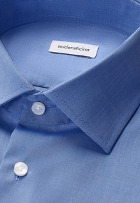 Jasnoniebieska koszula z kołnierzykiem typu spread i białymi guzikami, z białą metką z nazwą marki "seidensticker" wewnątrz kołnierzyka.