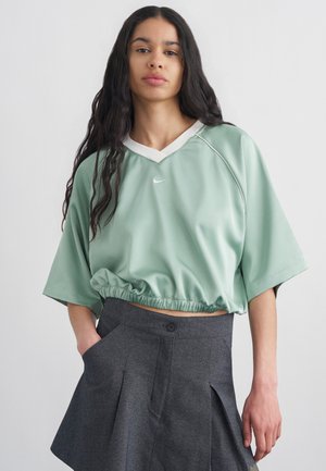 Femeie cu păr lung și închis la culoare, purtând un tricou scurt Nike larg de culoare verde mentă și o fustă plisată gri închis, stând în fața unui fundal simplu deschis.