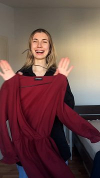 Χαμογελαστή γυναίκα κρατά ένα μακρυμάνικο μπορντό φόρεμα σε κρεμάστρα σε υπνοδωμάτιο με κρεβάτι και ουδέτερους τοίχους στο φόντο.