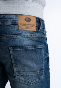 Granatowe dżinsy z denimu z skórzaną łatą z tekstem, tylnymi kieszeniami i lekko wypłowiałą fakturą. Pas jest szary, co odsłania koszulę spod spodu.