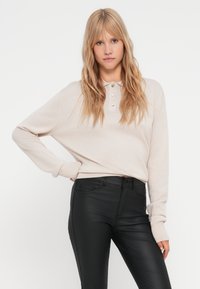 Bež majica s dugim rukavima s tri gumba na izrezu, uparena s crnim uskim hlačama. Glatka pletena tekstura s rebrastim manžetama.