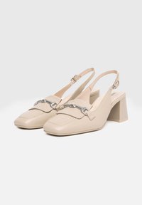 Scarpe slingback beige con punta quadrata, in materiale di pelle, accentate da una catena argentata e con tacco a blocco. Texture liscia e design minimalista.