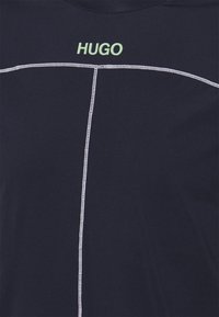 Námořnická modrá tričko s bílým prošíváním, které obrysuje geometrický design; na límci se nachází "HUGO" ve světle zelené barvě. Měkká struktura látky.