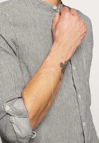 Gestreiftes Hemd mit Knöpfen in Schwarz und Weiß; verfügt über einen Stehkragen und hochgekrempelte Ärmel; der Stoff wirkt strukturiert und leicht.