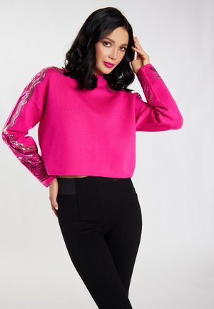 Fuchsia Cropped-Pullover mit hohem Kragen, mit glänzenden Wellenmustern auf den Ärmeln. Kombiniert mit eng anliegenden schwarzen Hosen.