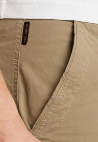 Beige katoenen broek met een gladde textuur, voorzien van een zijzak en een zwarte label met de tekst "PINE LEGEND" gestikt nabij de tailleband.