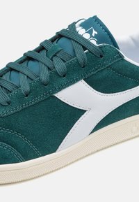 Sneaker verde in suede con dettagli in pelle bianca, lacci piatti e suola in gomma testurizzata; presenta una linguetta imbottita con stampa del logo.