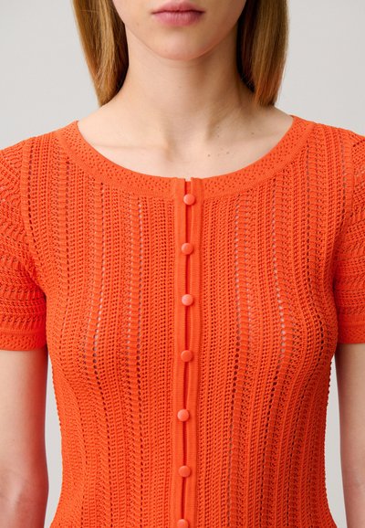 Primer plano de una persona que lleva una camiseta de punto de manga corta de color naranja brillante con un escote redondo y cierre delantero con botones verticales.