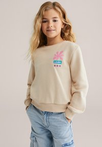 Jauna meitene ar garām blondām matēm, uzvilkusi bezrūpīgu bēšu kreklu ar rozā un zilām apdrukām, kā arī gaišzilus džinsu bikses ar kabatām, ar rokām kabatās.