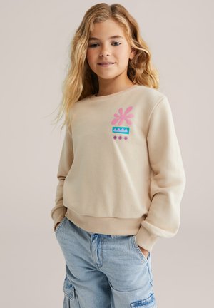 Mlada deklica z dolgimi blond lasmi, oblečena v bež pulover s pink in modrimi vzorci ter svetle modre jeans cargo hlače, z rokama v žepih.