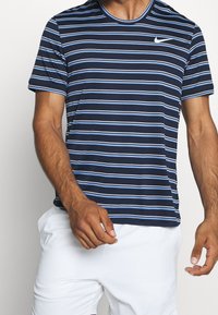 Muž v námořnickým modrém pruhovaném tričku Nike a bílých šortkách, stojící na jednobarevném světlém pozadí.