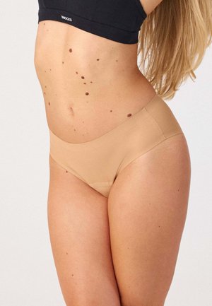 Frau trägt beige nahtlose Unterwäsche und ein schwarzes bauchfreies Top, zeigt den Oberkörper und obere Oberschenkel mit sichtbaren Muttermalen auf der Haut.