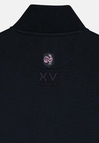 Polo noir avec un col haut, arborant un emblème de coq rose et un texte en relief "XV FRANCE" sous le blason. Texture de tissu subtile.