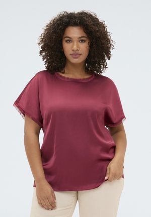 Vrouw met krullend haar die een losse, korte mouwen, kastanjebruine satijnen blouse en lichtbeige broek draagt tegen een effen lichte achtergrond.