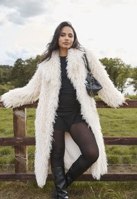 Biały, puszysty, długi płaszcz o wełnistej teksturze, noszony na czarnej sukience i czarnych rajstopach, w połączeniu z czarnymi, wysokimi kozakami i małą torebką.