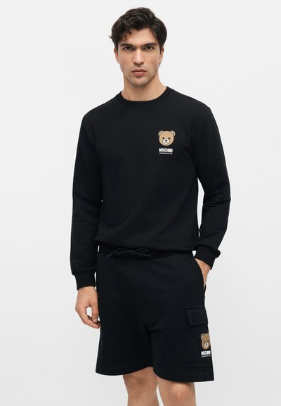 Άνδρας που φοράει μαύρο μπλουζάκι με μακριά μανίκια και σορτς με λογότυπα αρκούδας και το κείμενο "Moschino Underbear" στο στήθος και στην τσέπη του ποδιού.