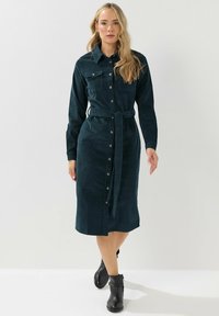 Abito camicia in velluto a coste color teal con maniche lunghe, apertura a bottoni frontale, due tasche sul petto e vita legata. Completato con stivali neri all'altezza della caviglia.