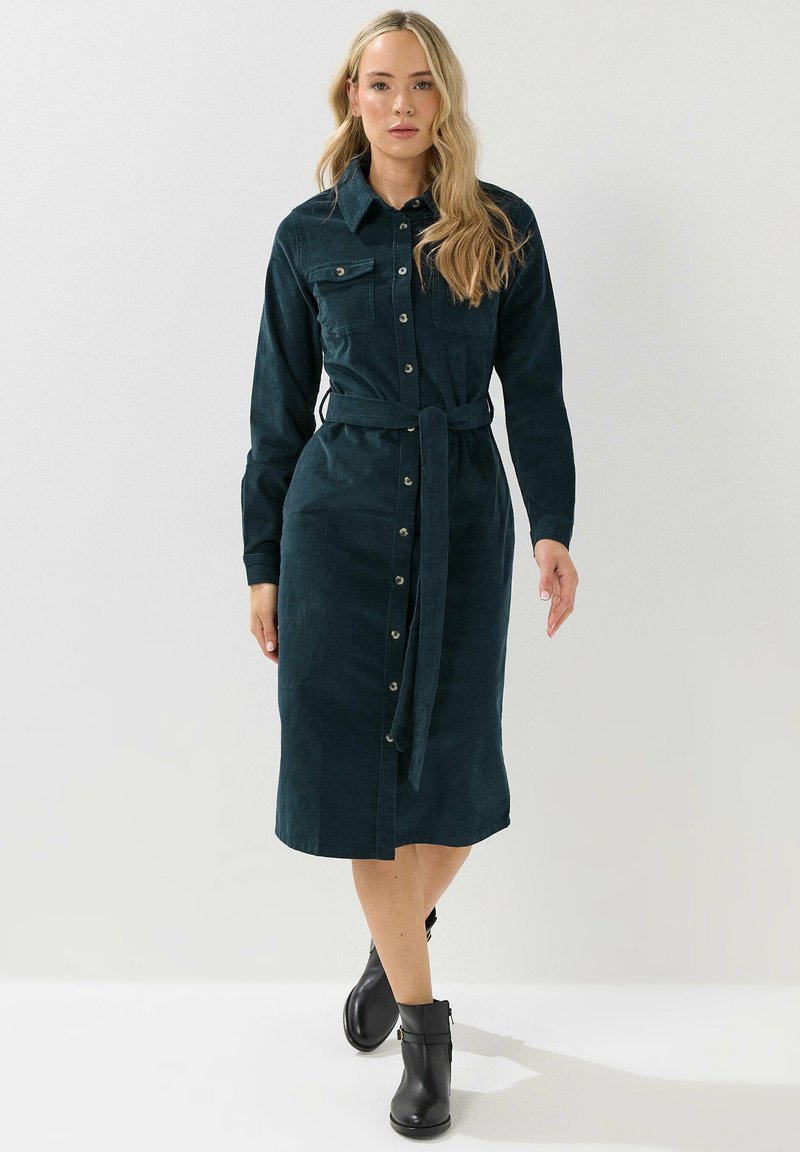 Abito camicia in velluto a coste color teal con maniche lunghe, apertura a bottoni frontale, due tasche sul petto e vita legata. Completato con stivali neri all'altezza della caviglia.
