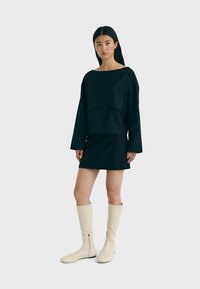 Schwarzes, übergroßes Langarmoberteil mit einem runden Ausschnitt und einem passenden schwarzen Minirock. Das Model trägt kniehohe cremefarbene Stiefel mit einem seitlichen Reißverschluss.