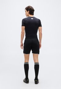  Νέος άνδρας που φοράει μαύρο αθλητικό μπλουζάκι, σορτς, ριγέ κάλτσες μέχρι το γόνατο και παπούτσια ποδοσφαίρου, στέκεται με την πλάτη προς την κάμερα σε λευκό φόντο.