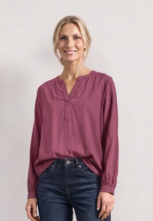 Femme blonde souriante, portant un chemisier prune à manches longues, rentré dans un jean bleu foncé, debout devant un fond clair uni.