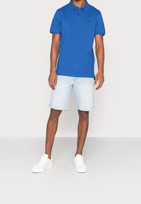 Modrá polo tričko s límečkem a černým logem, spárované s světlými denimovými šortkami a bílými teniskami. Hladká tkanina a dobře padnoucí střih.