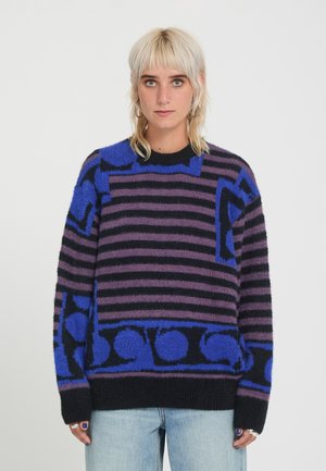 Jeune personne aux cheveux blonds clairs porte un pull rayé et à motifs noir, violet et bleu avec un jean bleu clair sur fond uni.