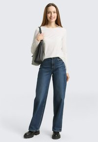 Witte blouse met lange mouwen, blauwe wijde jeans en zwarte laarzen met dikke zolen. Accessoires omvatten een zwarte handtas. Lichte achtergrond.