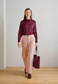 Žena hoda unutra noseći burgundsku satensku bluzu, svijetloružičaste hlače širokih nogavica, burgundske mokasine i nosi odgovarajuću burgundsku torbu.