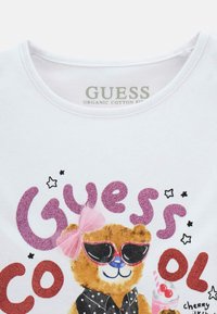 Biała koszulka z organicznej bawełny z grafiką niedźwiedzia w okularach przeciwsłonecznych, różową kokardą oraz kolorowym, brokatowym napisem "Guess Cool".