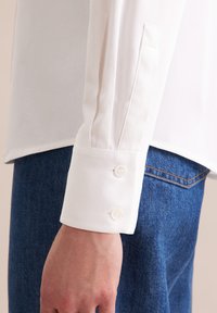 Biała koszula z długim rękawem, z guzikowym mankietem i detalami plisu. Materiał wydaje się gładki; noszona z niebieskim dżinsem.