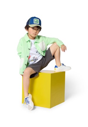 Mintgroene spijkerjas met witte zakken, witte grafische T-shirt, grijze korte broek, witte en blauwe sneakers, zittend op een gele kubus.