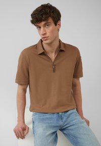 Braunes Polohemd mit Reißverschluss aus strukturiertem Material. Kurze Ärmel und klassischer Kragen. Kombiniert mit hellblauen Jeans.