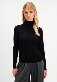 Blondynka z prostymi włosami nosi czarny sweter z golfem z złotymi guzikami na rękawach oraz szare szerokie spodnie w kant.