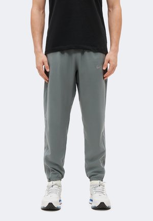 Férfi szürke Nike melegítőnadrágban és fehér Nike tornacipőben, világos egyszínű háttér előtt áll, kezei laza tartásban oldalt.