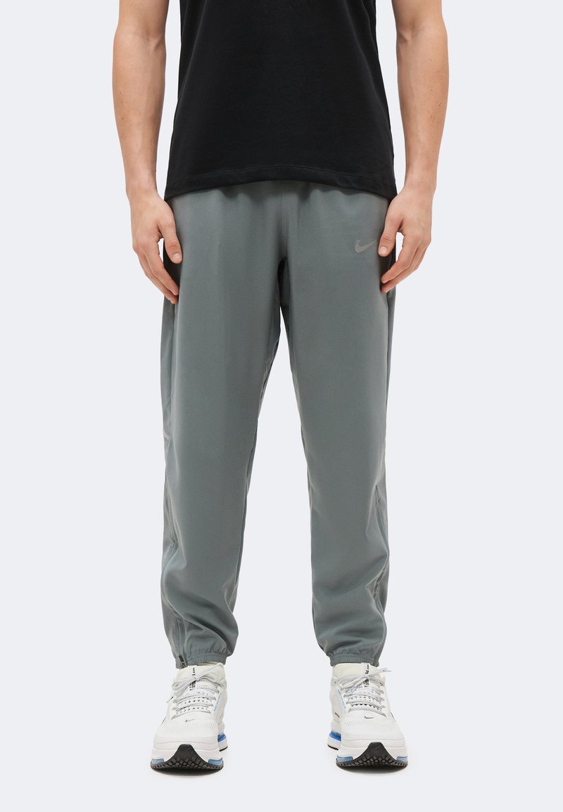 Άνδρας που φοράει γκρι παντελόνι τζόγκινγκ Nike και λευκά παπούτσια Nike, στέκεται μπροστά σε μια απλή φωτεινή επιφάνεια με τα χέρια χαλαρά δίπλα στο σώμα.