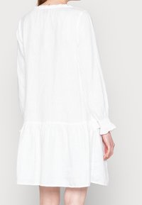 Vestido blanco de lino con mangas largas, escote con volantes y dobladillo en capas. La tela es ligera y de textura suave.