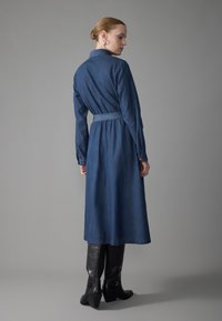 Jeansklänning med långa ärmar, åtsittande midja och full kjol. Mörkblå färg, synliga knappar på ärmarna, bärs med knähöga svarta stövlar.