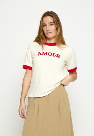 Žena s dugom kosom, odjevena u bijelu majicu s crvenim obrubom i tekstom "AMOUR", uparenu s bež plisiranom suknjom, stoji na jednobojnoj pozadini.