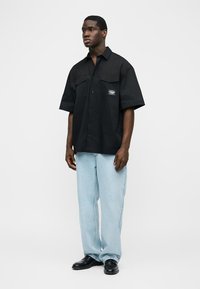 Muž v černé krátkosleevé košili na knoflíky a světle modrých džínách stojící před jednobarevným pozadím.