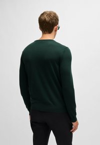 Ciemnozielony sweter dziewiarski z długimi rękawami i okrągłym dekoltem; gładka tekstura i dopasowany krój, w połączeniu z ciemnymi spodniami.