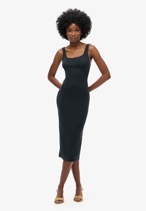 Kobieta z kręconymi włosami, ubrana w czarną, bezrękawnikową sukienkę midi i beżowe, odkryte buty na obcasie, stojąca z rękami za plecami na białym tle.