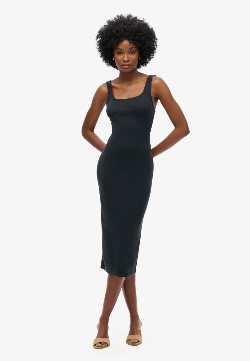 Kvinde med krøllet hår iført en sort midi-kjole uden ærmer og beige åbne tå-sandaler, stående med hænderne bag ryggen på en hvid baggrund.