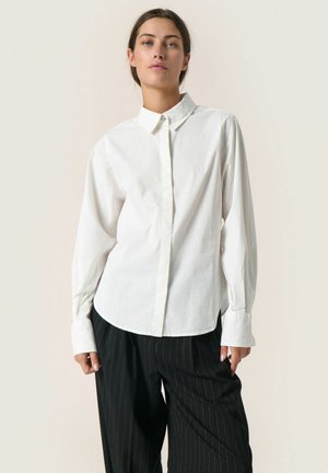 White long-sleeve shirt with a classic collar, button front, and relaxed fit. Paired with pinstriped black pants for contrast.