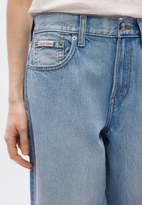 Ανοιχτό μπλε τζιν μεσαίας μέσης, ορατές ραφές, ορθογώνια μπροστινή τσέπη και ετικέτα Calvin Klein στην πίσω τσέπη.