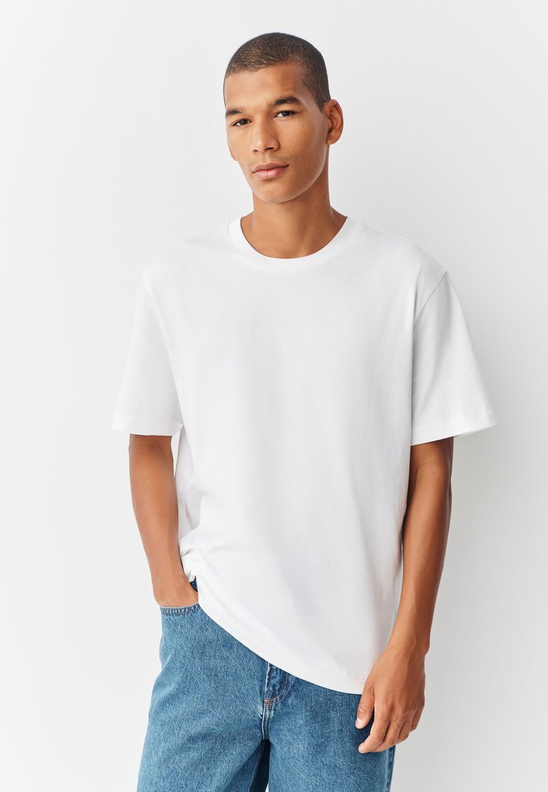 Tricou alb din bumbac, cu decolteu rotund și mâneci scurte, asortat cu pantaloni scurți din denim albastru. Tricoul are o croială lejeră și o textură fină.