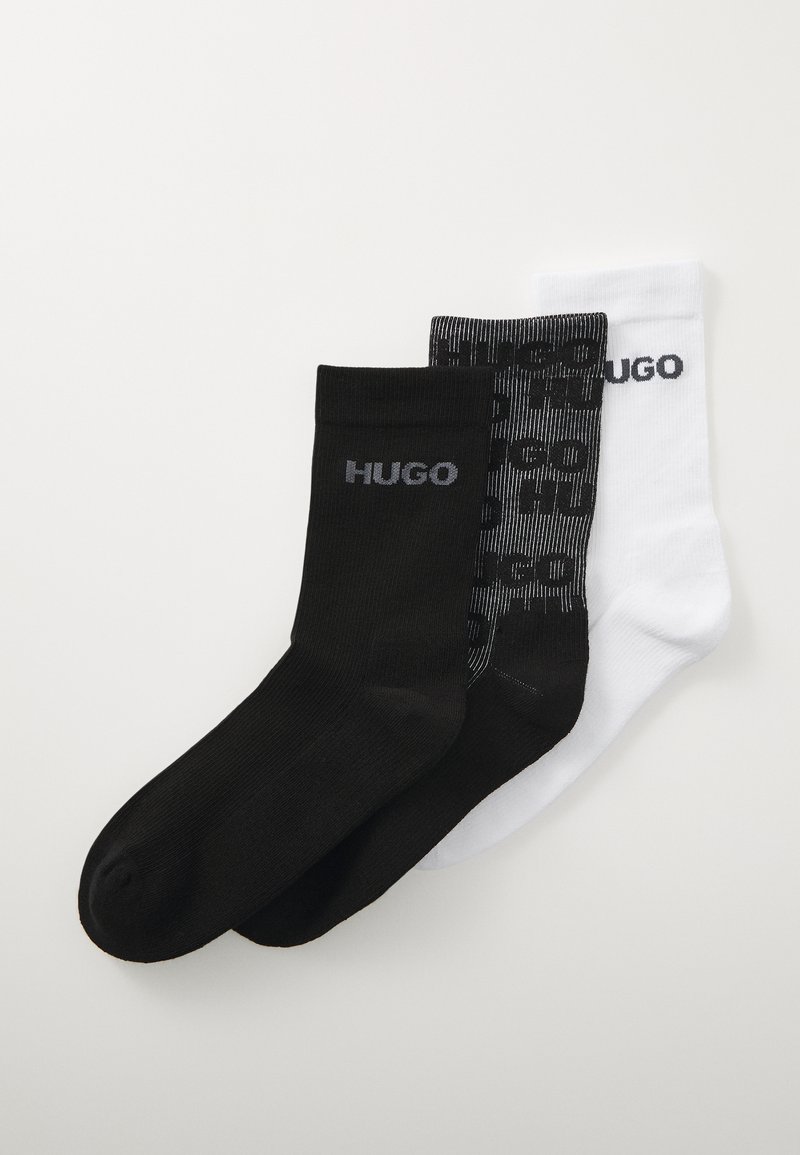 Tres pares de calcetines: uno negro sólido, uno blanco sólido y uno con un patrón negro con un logo gris. Todos son de longitud media.