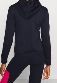 Sudadera con capucha azul marino con cordón ajustable, puños ceñidos y dobladillo. Se lleva con leggings oscuros a juego que tienen un detalle rosa en la pierna.